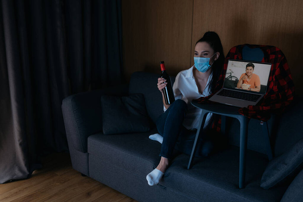 junge Frau in medizinischer Maske mit einer Flasche Wein auf dem Sofa neben dem Laptop sitzend mit einem lächelnden asiatischen Freund auf dem Bildschirm - Foto, Bild