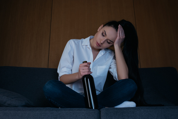 depressed woman touching head while sitting on sofa with bottle of wine - Foto, Imagem
