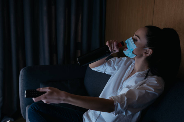 mujer deprimida en máscara médica con agujero beber vino de la botella mientras se utiliza el control remoto de televisión
 - Foto, Imagen