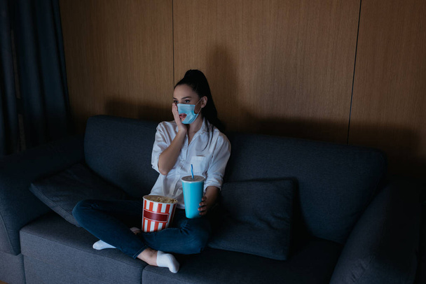hoge hoek uitzicht van bezorgde jonge vrouw in medisch masker met gat kijken tv met frisdrank en popcorn thuis - Foto, afbeelding