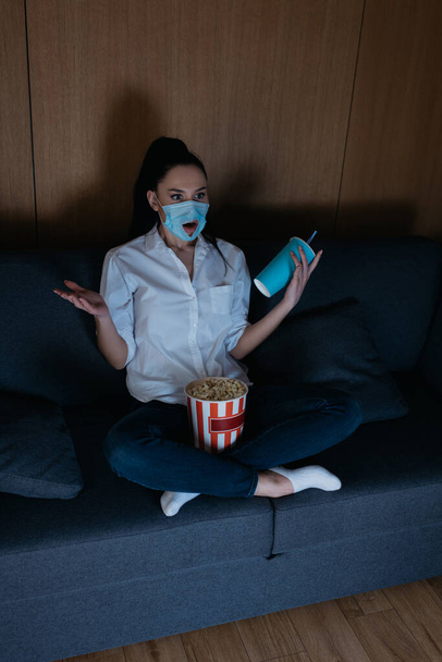 άποψη υψηλής γωνίας του σοκαρισμένη νεαρή γυναίκα σε ιατρική μάσκα με τρύπα βλέποντας τηλεόραση με ποπ κορν και σόδα στο σπίτι - Φωτογραφία, εικόνα