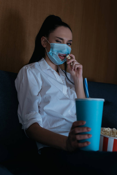 επιλεκτική εστίαση της νεαρής γυναίκας στην ιατρική μάσκα με τρύπα κλείσιμο του ματιού στην κάμερα, ενώ κρατώντας σόδα και τρώγοντας ποπ κορν - Φωτογραφία, εικόνα