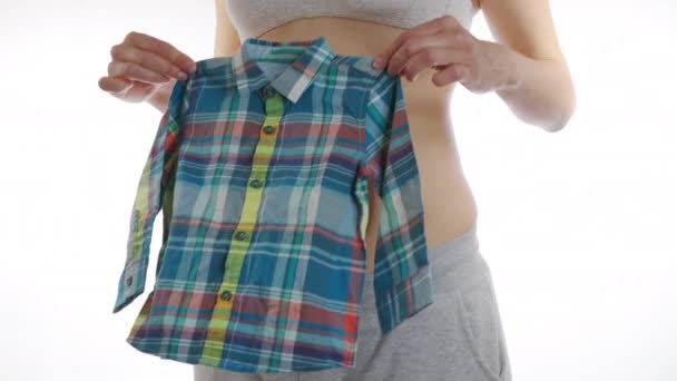 Szczęśliwa kobieta w ciąży stosuje ubrania dla niemowląt do brzucha dziecka. Macierzyństwo, ciąża, koncepcja ubrań ludzi i dzieci. Zbliżenie, 6k. - Materiał filmowy, wideo