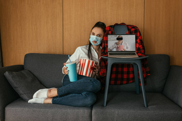 jovem mulher em máscara médica com buraco segurando pipoca e refrigerante perto de laptop com namorado no fone de ouvido vr na tela
 - Foto, Imagem