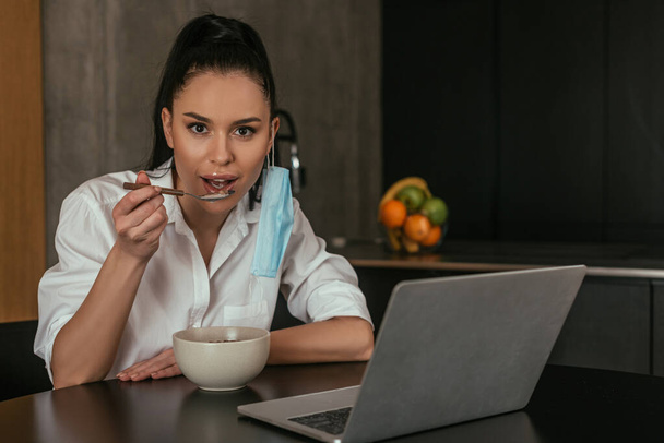 young woman having breakfast and looking at camera near laptop in kitchen - Photo, Image