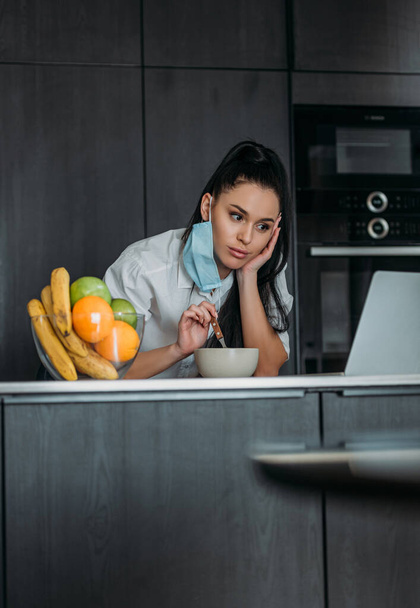 pensive woman holding spoon while looking at laptop near bowl with fruits in kitchen - Zdjęcie, obraz