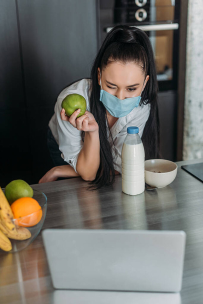 jeune femme dans le masque médical tenant pomme et clin d'oeil pendant l'appel vidéo dans la cuisine
 - Photo, image