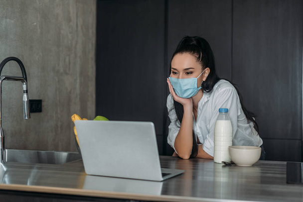 young woman in protective mask looking at laptop near bottle of milk in kitchen - Фото, зображення