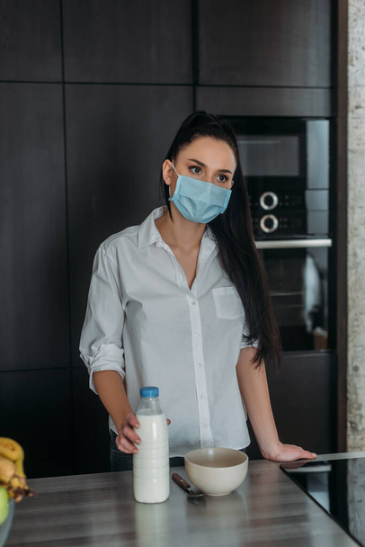 θλιμμένη γυναίκα με ιατρική μάσκα κοιτάζει αλλού ενώ αγγίζει μπουκάλι γάλα στην κουζίνα - Φωτογραφία, εικόνα