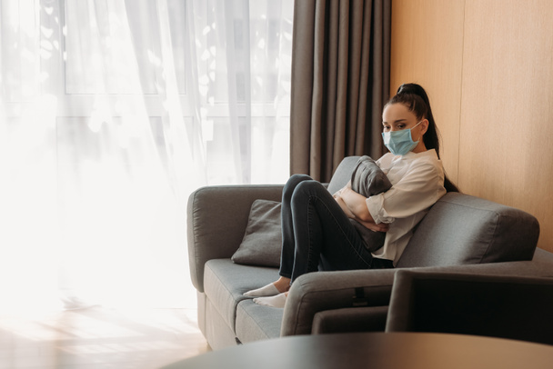 upset young woman in protective mask sitting on sofa and hugging pillow - Foto, imagen