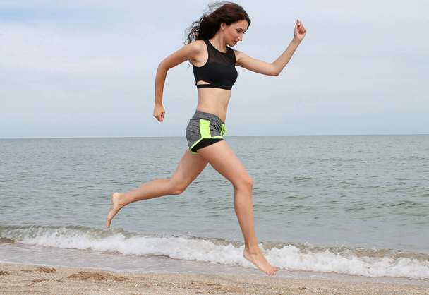 Rennende jonge brunette vrouw. Vrouwelijk hardlopen tijdens outdoor training op het strand. Slank fitnessmodel buiten - Foto, afbeelding