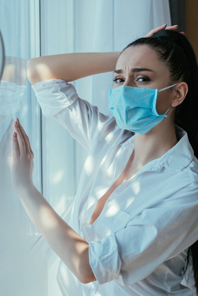 depressed girl in medical mask touching head while standing near window at home - Photo, Image