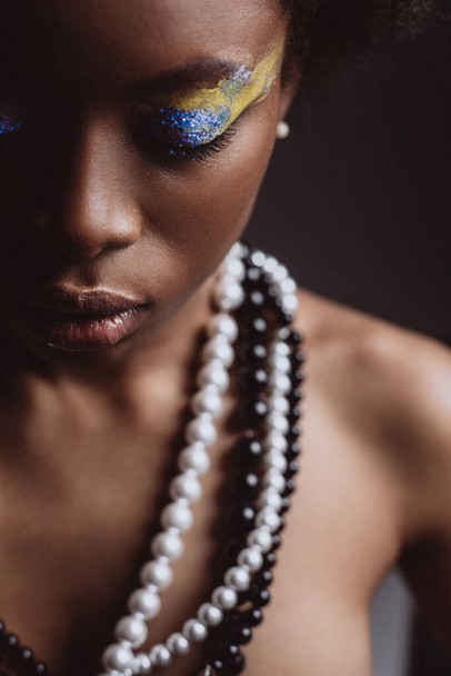 Emotional close up portrait with closed eyes of an African in jewelry on black background with creative make-up . - Photo, Image