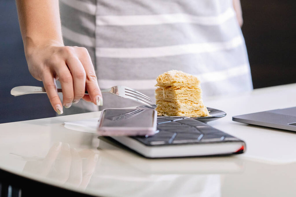 Female hand holds a puff cake in a plate on the table. Blurry diary cup of coffee, laptop and smartphone. Selected Focus. Coronavirus quarantine homework, social distance, private entrepreneur. - Photo, image