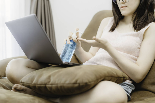Młoda kobieta siedzieć na kanapie i za pomocą komputera laptopa umieścić poduszkę i za pomocą algoholu żel lub dezynfektor do mycia rąk. Praca koncepcyjna z domu i dystans społeczny z covid-19 - Zdjęcie, obraz
