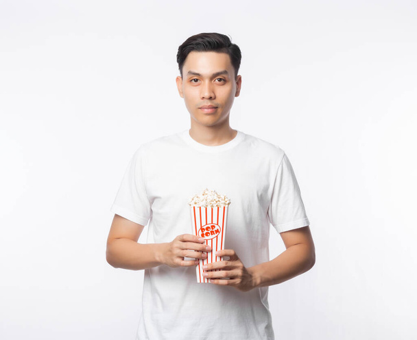 Joven asiático hombre en blanco camiseta sosteniendo palomitas de maíz y mirando a cámara aislada en blanco fondo
. - Foto, Imagen