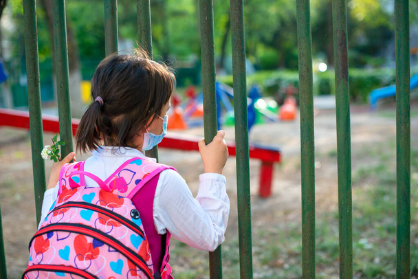 Little school girl sitting next to school fence waiting for going back to clases after pandemic outbreak - Photo, Image