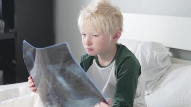 Ένα όμορφο ξανθό αγόρι μελετάει μια φωτογραφία των πνευμόνων του στο νοσοκομείο.. - Πλάνα, βίντεο