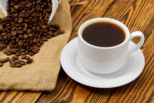 Біла чашка кави з ароматним кавовим напоєм на коричневому дерев'яному столі. Насіння кави на старовинному дерев'яному столі з чашкою кавового напою
 - Фото, зображення