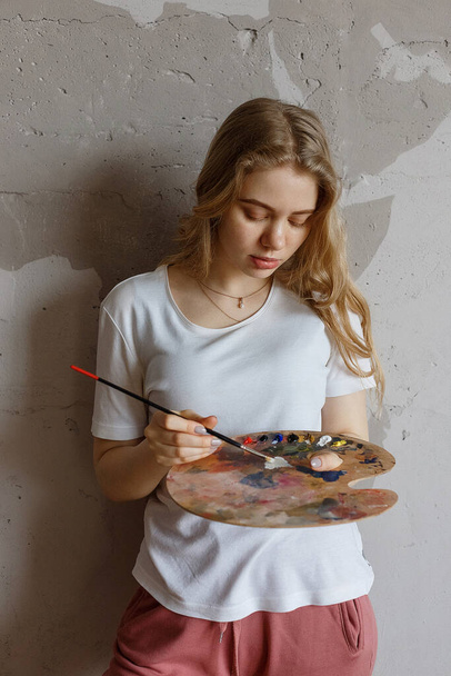 Young pensive pretty blonde girl with brush and color palette. Art, creativity, hobby concept, painting process during COVID-19 pandemic self isolation quarantine. stay home, 2019-nCoV prevention - Fotoğraf, Görsel