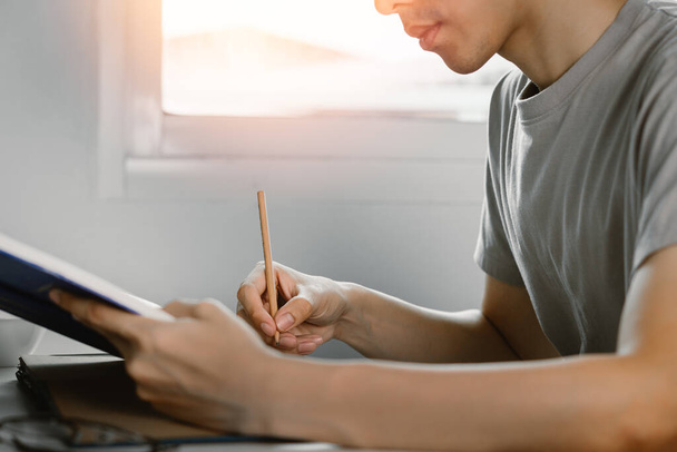 Νεαρός άνδρας διαβάζει βιβλίο και γράφει σε σημείωμα στο γραφείο εργασίας στον ελεύθερο χρόνο από την εργασία στο σπίτι, Γνώση και μάθηση έννοια. - Φωτογραφία, εικόνα