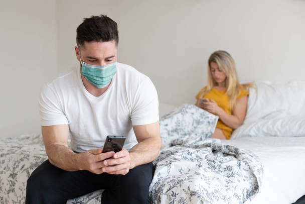 Couple under Covid-19 quarantine using their phones at home. Man wearing face mask texting on smartphone. - Foto, Bild