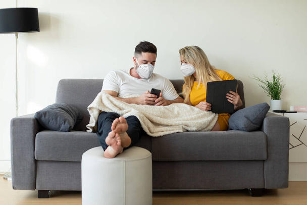 Couple wearing face mask using digital devices under Covid-19 quarantine. Digital leisure at home under coronavirus worldwide crisis, - Photo, Image
