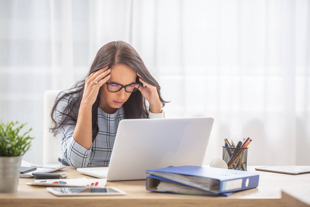 Kobieta z migreną trzymającą głowę w rękach z zamkniętymi oczami przy biurku, przed komputerem. - Zdjęcie, obraz