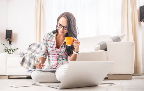 Woman having a tea, sitting on a floor in an apartment, writing down notes, opened laptop in front of her. Home office concept. - Foto, Imagen
