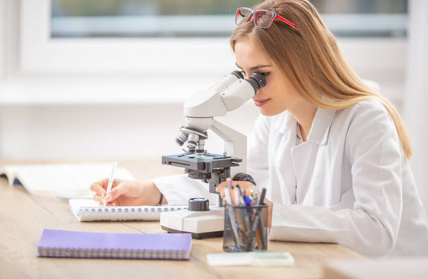 Beautiful young student girl researcher looking into a microscope, writing down notes. - Φωτογραφία, εικόνα