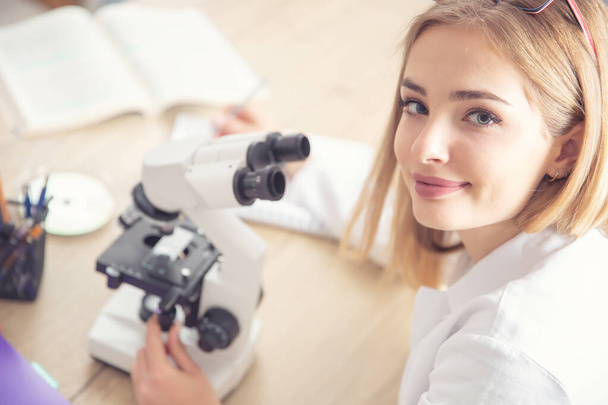 Piękna młoda kobieta patrząc do kamery podczas pracy z mikroskopem, zapisując notatki. - Zdjęcie, obraz