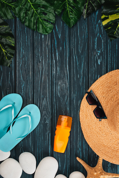 Top view of sunscreen, flip flops near straw hat and pebbles on dark wooden surface - Fotografie, Obrázek