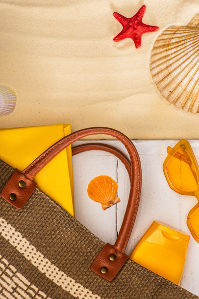 Bovenaanzicht van tas, zonnebrandcrème en boek op witte houten planken bij schelpen op zand - Foto, afbeelding
