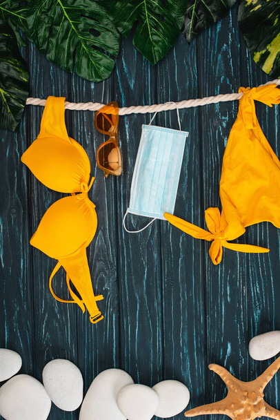 Top view of yellow swimsuit near medical mask, sunglasses and leaves on dark wooden surface - 写真・画像