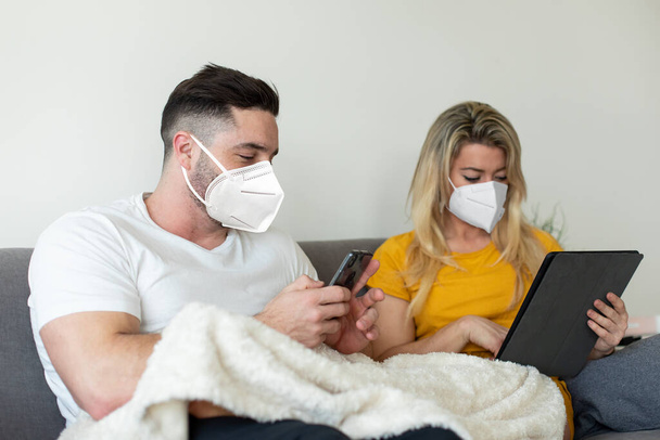 Couple wearing face mask using digital devices under Covid-19 quarantine. Digital leisure at home under coronavirus worldwide crisis, - Foto, afbeelding