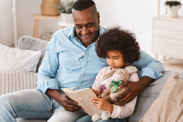 Leyendo con el abuelo. Hombre y nieta afroamericanos mayores con libro cautivador en casa
 - Foto, Imagen