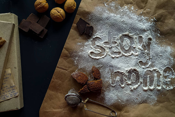 De inscriptie blijft thuis gemaakt van witte tarwemeel. Verspreide koekjes, noten, chocolade op een donkere achtergrond, handgeschreven meelwoord Blijf thuis. - Foto, afbeelding
