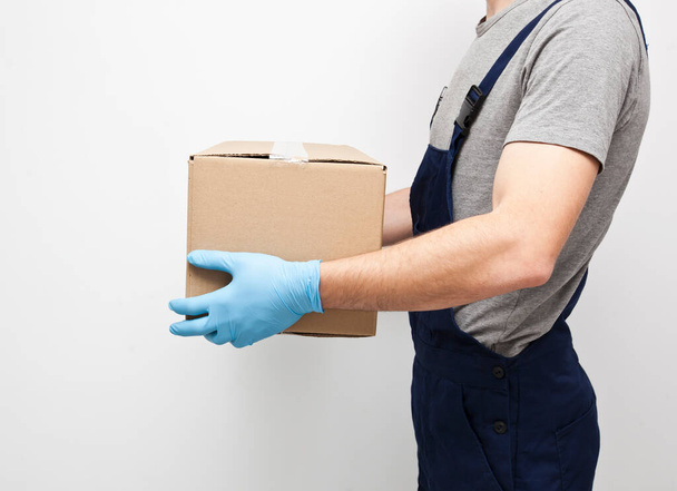 The courier is holding the carton box in his hand wearing  protective latex gloves on the gray background - Fotoğraf, Görsel