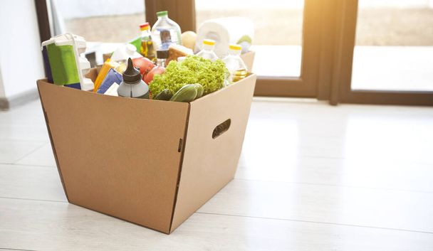 La caja ecológica de cartón completo con productos de la tienda de comestibles en el suelo en casa cerca de la puerta - Foto, Imagen