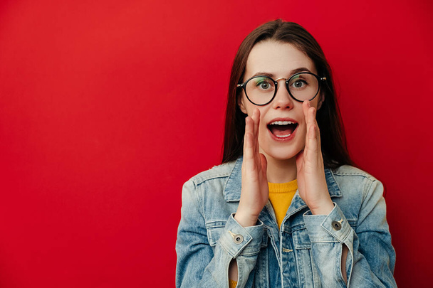 Portrait of young brunette woman screams with excitement, keeps hands near mouth, wears denim jacket and eyeglasses, posing isolated on red background. People lifestyle concept. Mock up copy space. - Foto, afbeelding