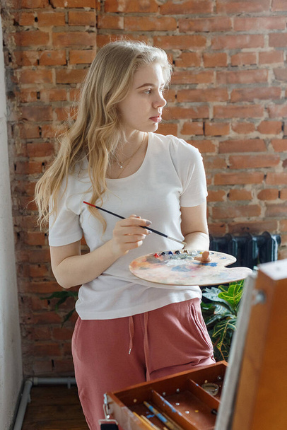 stay home: young pretty blonde girl with brush and palette standing near easel drawing picture. Art, creativity, hobby concept, painting process during COVID-19 pandemic self isolation quarantine - Photo, Image