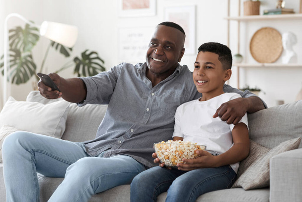 Ευτυχισμένος Αφροαμερικανός παππούς βλέποντας τηλεόραση με τον εγγονό στο σπίτι μαζί - Φωτογραφία, εικόνα