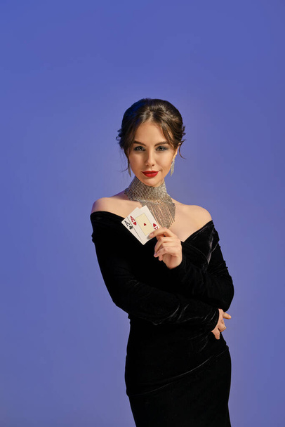 Barna lány, csupasz vállakkal, fekete ruhában és csillogó ékszerekkel. Két kártyajátékot mutat, lila alapon pózol. Póker, kaszinó. Közelkép - Fotó, kép