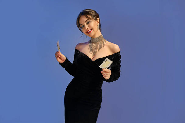 Brunette girl with bare shoulders, in black dress and jewelry. Smiling, showing two playing cards posing on purple background. Poker, casino. Close-up - Photo, Image