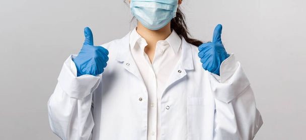 Doctors, infectionist, research and covid19 concept. Satisfied young asian female doctor receive good results on studying virus, discover vaccine, show thumb-up, wear face mask and gloves - Photo, Image