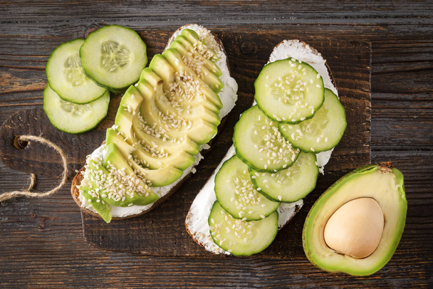 2 sandwiches with cheese, avocado and cucumber on a dark wooden background, Breakfast, snack, pieces of green cucumber, half of fresh avocado - Фото, зображення