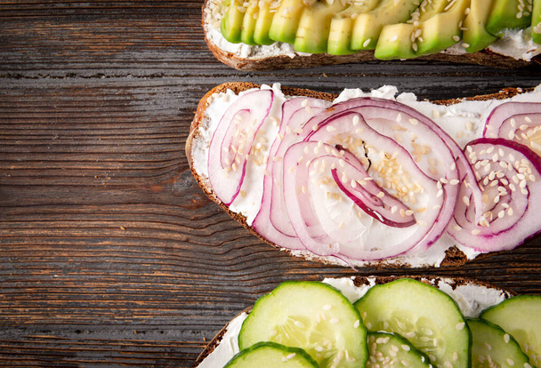 3 homemade  sandwiches with cheese, avocado,  cucumber , onion and sesame on a dark wooden background, Breakfast, snack,  top view, copy space, - 写真・画像