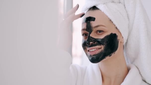 Pretty woman with wrapped hair in a towel is applying black mask on face - Filmagem, Vídeo