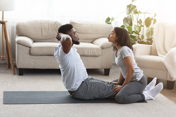 Siyah çift evde birlikte egzersiz yapıyor, kadın erkek arkadaşına karın kası egzersizlerinde yardım ediyor. - Fotoğraf, Görsel