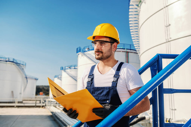 Young hardworking Caucasian unshaven refinery worker in working clothes with helmet on head and protective glasses holding folder with documents. - Photo, Image